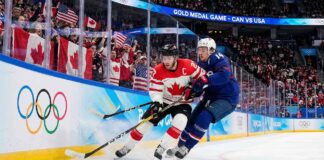 Team Canada captain Connor McDavid battles intensely with Team USA defenseman Charlie McAvoy for a loose puck along the arena boards during a men's Olympic gold medal ice hockey game. Fans wave Canadian and American flags in the background.