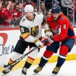 Vegas Golden Knights forward Jack Eichel (9), wearing a white away jersey, and Washington Capitals captain Alex Ovechkin (8), wearing a red home jersey, engage in a physical battle with their sticks for the puck along the rink boards in front of cheering fans at Capital One Arena.