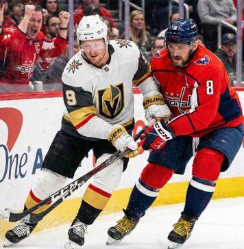 Vegas Golden Knights forward Jack Eichel (9), wearing a white away jersey, and Washington Capitals captain Alex Ovechkin (8), wearing a red home jersey, engage in a physical battle with their sticks for the puck along the rink boards in front of cheering fans at Capital One Arena.