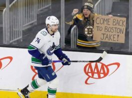 Vancouver Canucks forward Jake DeBrusk (74) skates along the rink boards while a female fan in a Boston Bruins jersey and hat behind the glass excitedly holds up a handmade cardboard sign that reads, "COME BACK TO BOSTON JAKE!".