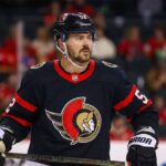 A photo illustration showing NHL defenseman MacKenzie Weegar on the ice, wearing a black Ottawa Senators home jersey with the centurion logo and number 52, holding his hockey stick.