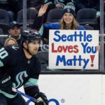 Seattle Kraken center Matty Beniers (#10) skates with the puck during a game. In the background behind the glass, a smiling female fan wearing Kraken gear holds up a handmade sign that reads "Seattle Loves You Matty!" in blue and red letters.