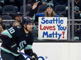 Seattle Kraken center Matty Beniers (#10) skates with the puck during a game. In the background behind the glass, a smiling female fan wearing Kraken gear holds up a handmade sign that reads "Seattle Loves You Matty!" in blue and red letters.