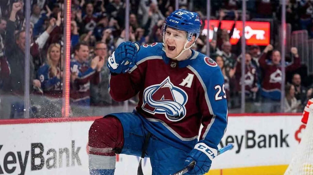 Colorado Avalanche forward Nathan MacKinnon, wearing the blue and burgundy alternate jersey with the number 29, celebrates a goal by yelling and pumping his fist while on one knee on the ice. Ice spray is visible around his leg, and fans are cheering in the background behind the glass.