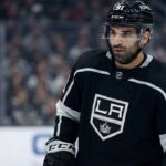 Los Angeles Kings center Nazem Kadri, wearing his black number 91 home jersey and helmet with a clear visor, stands on the ice looking towards the left side of the frame with a serious expression. The background shows blurred spectators in the arena stands.