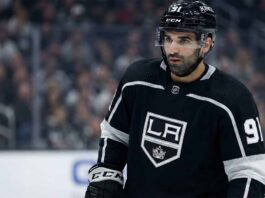 Los Angeles Kings center Nazem Kadri, wearing his black number 91 home jersey and helmet with a clear visor, stands on the ice looking towards the left side of the frame with a serious expression. The background shows blurred spectators in the arena stands.