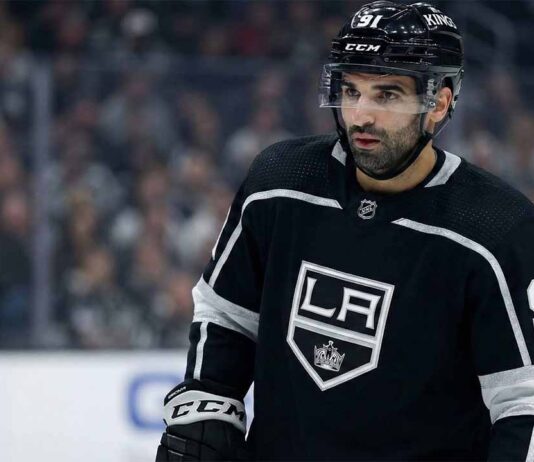 Los Angeles Kings center Nazem Kadri, wearing his black number 91 home jersey and helmet with a clear visor, stands on the ice looking towards the left side of the frame with a serious expression. The background shows blurred spectators in the arena stands.