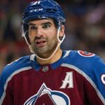 Missing Piece? Why the Colorado Avalanche Must Trade for Nazem Kadri A close-up photograph of Colorado Avalanche forward Nazem Kadri (#91) wearing a blue helmet with a visor and a burgundy and blue jersey featuring the alternate captain 'A' patch, looking intently towards the side during an NHL game.