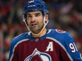 Missing Piece? Why the Colorado Avalanche Must Trade for Nazem Kadri A close-up photograph of Colorado Avalanche forward Nazem Kadri (#91) wearing a blue helmet with a visor and a burgundy and blue jersey featuring the alternate captain 'A' patch, looking intently towards the side during an NHL game.