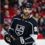 Nazem Kadri depicted wearing a black Los Angeles Kings number 91 hockey jersey, holding his stick and looking towards the right side of the frame against a blurred arena crowd background.