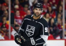 Nazem Kadri depicted wearing a black Los Angeles Kings number 91 hockey jersey, holding his stick and looking towards the right side of the frame against a blurred arena crowd background.
