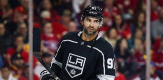 Nazem Kadri depicted wearing a black Los Angeles Kings number 91 hockey jersey, holding his stick and looking towards the right side of the frame against a blurred arena crowd background.