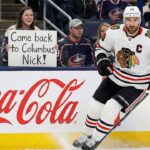 Chicago Blackhawks captain Nick Foligno (#17) skates near the boards during an NHL game. In the background, a female fan wearing a Columbus Blue Jackets jersey holds up a handwritten sign that reads, "Come back to Columbus Nick!". A large red Coca-Cola advertisement is visible on the dasher boards beneath the fan.