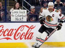 Chicago Blackhawks captain Nick Foligno (#17) skates near the boards during an NHL game. In the background, a female fan wearing a Columbus Blue Jackets jersey holds up a handwritten sign that reads, "Come back to Columbus Nick!". A large red Coca-Cola advertisement is visible on the dasher boards beneath the fan.