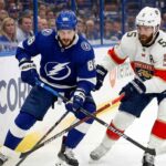 Tampa Bay Lightning forward Nikita Kucherov (86) in a blue jersey and Florida Panthers defenseman Aaron Ekblad (5) in a white jersey battle for the puck along the boards during an NHL game.