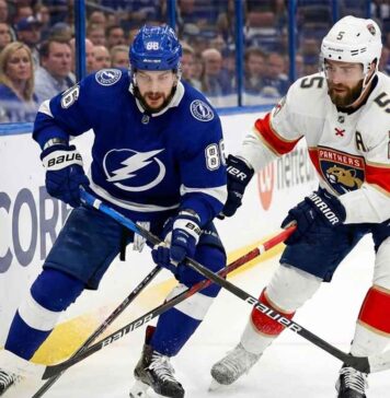 Tampa Bay Lightning forward Nikita Kucherov (86) in a blue jersey and Florida Panthers defenseman Aaron Ekblad (5) in a white jersey battle for the puck along the boards during an NHL game.