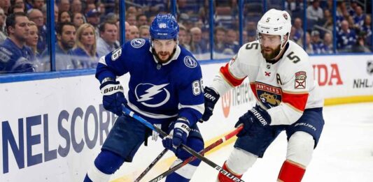 Tampa Bay Lightning forward Nikita Kucherov (86) in a blue jersey and Florida Panthers defenseman Aaron Ekblad (5) in a white jersey battle for the puck along the boards during an NHL game.
