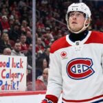Patrik Laine, wearing a white Montreal Canadiens away jersey and helmet, looks up while on the ice. Behind the glass boards, a young female fan wearing a Canadiens toque holds a handmade sign with blue and red lettering that reads, "Good luck in Calgary Patrik!".
