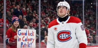 Patrik Laine, wearing a white Montreal Canadiens away jersey and helmet, looks up while on the ice. Behind the glass boards, a young female fan wearing a Canadiens toque holds a handmade sign with blue and red lettering that reads, "Good luck in Calgary Patrik!".
