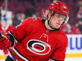 A close-up photograph of professional ice hockey player Patrik Laine wearing a red Carolina Hurricanes jersey with the team's logo, a red helmet with "HURRICANES" and number 92, and red gloves. He is holding a hockey stick and