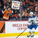 St. Louis Blues center Robert Thomas (#18) skates with the puck along the dasher boards at the Wells Fargo Center in Philadelphia. In the background, behind the glass, a female fan wearing Philadelphia Flyers gear holds up a handwritten sign with orange lettering that reads, "Come to Philly Robert!".
