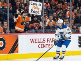 St. Louis Blues center Robert Thomas (#18) skates with the puck along the dasher boards at the Wells Fargo Center in Philadelphia. In the background, behind the glass, a female fan wearing Philadelphia Flyers gear holds up a handwritten sign with orange lettering that reads, "Come to Philly Robert!".