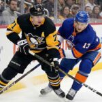 Pittsburgh Penguins center Evgeni Malkin (71) in a black road jersey and New York Islanders center Mathew Barzal (13) in a blue home jersey battle intensely for puck possession along the rink boards during an NHL game.