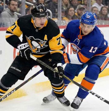 Pittsburgh Penguins center Evgeni Malkin (71) in a black road jersey and New York Islanders center Mathew Barzal (13) in a blue home jersey battle intensely for puck possession along the rink boards during an NHL game.