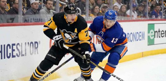 Pittsburgh Penguins center Evgeni Malkin (71) in a black road jersey and New York Islanders center Mathew Barzal (13) in a blue home jersey battle intensely for puck possession along the rink boards during an NHL game.