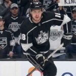 A visualization of hockey player Robert Thomas wearing a black No. 18 Los Angeles Kings jersey, skating intently on the ice while fans in casual clothing watch from behind the glass.