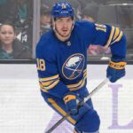 Robert Thomas wearing a blue Buffalo Sabres jersey with the number 18, blue helmet, blue gloves, and holding a hockey stick, is skating on the ice during a game. Spectators are visible in the background behind the glass boards.