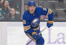 Robert Thomas wearing a blue Buffalo Sabres jersey with the number 18, blue helmet, blue gloves, and holding a hockey stick, is skating on the ice during a game. Spectators are visible in the background behind the glass boards.