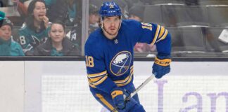 Robert Thomas wearing a blue Buffalo Sabres jersey with the number 18, blue helmet, blue gloves, and holding a hockey stick, is skating on the ice during a game. Spectators are visible in the background behind the glass boards.