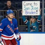 New York Rangers defenseman Braden Schneider skates past the boards where a female San Jose Sharks fan holds up a handwritten sign reading "Come to San Jose Braden!" amid trade rumors.