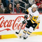 Pittsburgh Penguins captain Sidney Crosby (#87) is skating on the ice in his white road jersey, handling the puck near the boards which feature large advertisements for FanDuel Sportsbook and Coca-Cola.