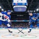 A face-off between Team Slovakia (white jersey, left) and Team Finland (blue jersey, right) ice hockey players during the preliminary round at the 2026 Winter Olympics in Milan. The arena is packed with fans, the Olympic rings are visible on the ice, and the center scoreboard above indicates a 1-1 tie.