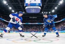 A face-off between Team Slovakia (white jersey, left) and Team Finland (blue jersey, right) ice hockey players during the preliminary round at the 2026 Winter Olympics in Milan. The arena is packed with fans, the Olympic rings are visible on the ice, and the center scoreboard above indicates a 1-1 tie.