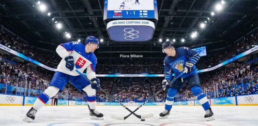 A face-off between Team Slovakia (white jersey, left) and Team Finland (blue jersey, right) ice hockey players during the preliminary round at the 2026 Winter Olympics in Milan. The arena is packed with fans, the Olympic rings are visible on the ice, and the center scoreboard above indicates a 1-1 tie.
