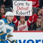 The Robert Thomas Price is Set: Can the Red Wings Pay It? St. Louis Blues center Robert Thomas (18) skates on the ice during a game, while a female fan in a Detroit Red Wings jersey holds up a sign in the background that reads "Come to Detroit Robert!".