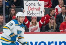 St. Louis Blues center Robert Thomas (18) skates on the ice during a game, while a female fan in a Detroit Red Wings jersey holds up a sign in the background that reads "Come to Detroit Robert!".