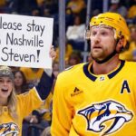 Nashville Predators alternate captain Steven Stamkos stands on the ice near the boards, looking upwards. Behind the protective glass, a smiling female fan wearing a Predators jersey and beanie holds up a handmade white sign with black text that reads, "Please stay in Nashville Steven!". The arena crowd is visible in the background.