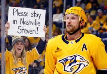 Nashville Predators alternate captain Steven Stamkos stands on the ice near the boards, looking upwards. Behind the protective glass, a smiling female fan wearing a Predators jersey and beanie holds up a handmade white sign with black text that reads, "Please stay in Nashville Steven!". The arena crowd is visible in the background.