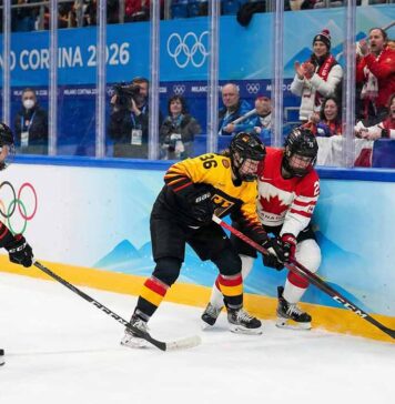 Team Canada's forward #27 and two Team Germany defenders #29 and #36 battle for control of the puck along the boards during the women's ice hockey quarterfinal match at the Milano Cortina 2026 Winter Olympics. The rink boards feature Olympic branding and spectators are cheering in the background.