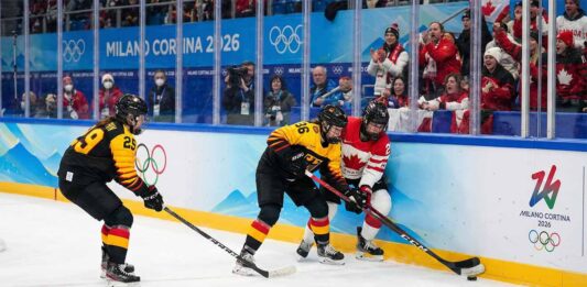 Team Canada's forward #27 and two Team Germany defenders #29 and #36 battle for control of the puck along the boards during the women's ice hockey quarterfinal match at the Milano Cortina 2026 Winter Olympics. The rink boards feature Olympic branding and spectators are cheering in the background.