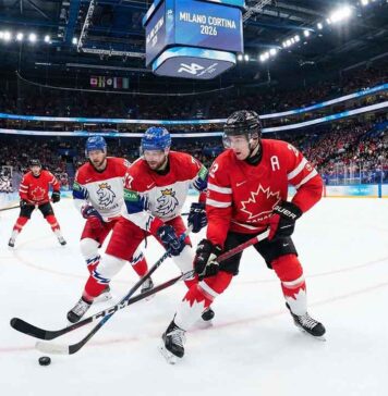 Action photograph of a men's ice hockey game during the Milano Cortina 2026 Winter Olympics. A Team Canada player in a red jersey is battling two Team Czechia players in white jerseys for puck possession near the center ice faceoff circle. The arena is packed with fans, and the scoreboard overhead displays the "MILANO CORTINA 2026" branding.