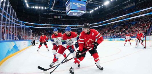 Action photograph of a men's ice hockey game during the Milano Cortina 2026 Winter Olympics. A Team Canada player in a red jersey is battling two Team Czechia players in white jerseys for puck possession near the center ice faceoff circle. The arena is packed with fans, and the scoreboard overhead displays the "MILANO CORTINA 2026" branding.