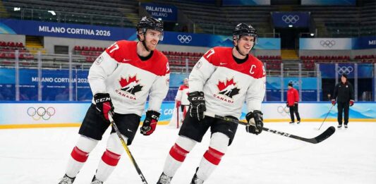 Connor McDavid (left) and captain Sidney Crosby (right) of Team Canada, both smiling and holding hockey sticks, skate together during a practice session at the Milano Cortina 2026 Winter Olympics arena.