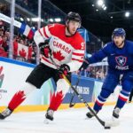 Team Canada captain Connor McDavid (#97) skates with the puck, shielding it from a Team France defender during a men's preliminary round ice hockey game at the Milano Cortina 2026 Winter Olympics. The arena boards feature official Olympic branding, and fans waving Canadian and French flags fill the stands.