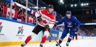Team Canada captain Connor McDavid (#97) skates with the puck, shielding it from a Team France defender during a men's preliminary round ice hockey game at the Milano Cortina 2026 Winter Olympics. The arena boards feature official Olympic branding, and fans waving Canadian and French flags fill the stands.