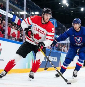 Team Canada captain Connor McDavid (#97) skates with the puck, shielding it from a Team France defender during a men's preliminary round ice hockey game at the Milano Cortina 2026 Winter Olympics. The arena boards feature official Olympic branding, and fans waving Canadian and French flags fill the stands.